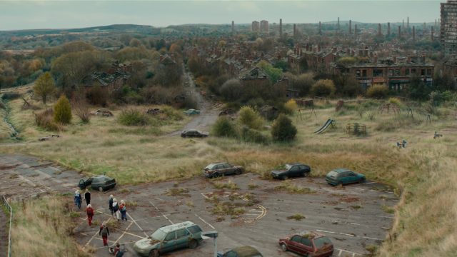People walking by cars with a derelict building in the background